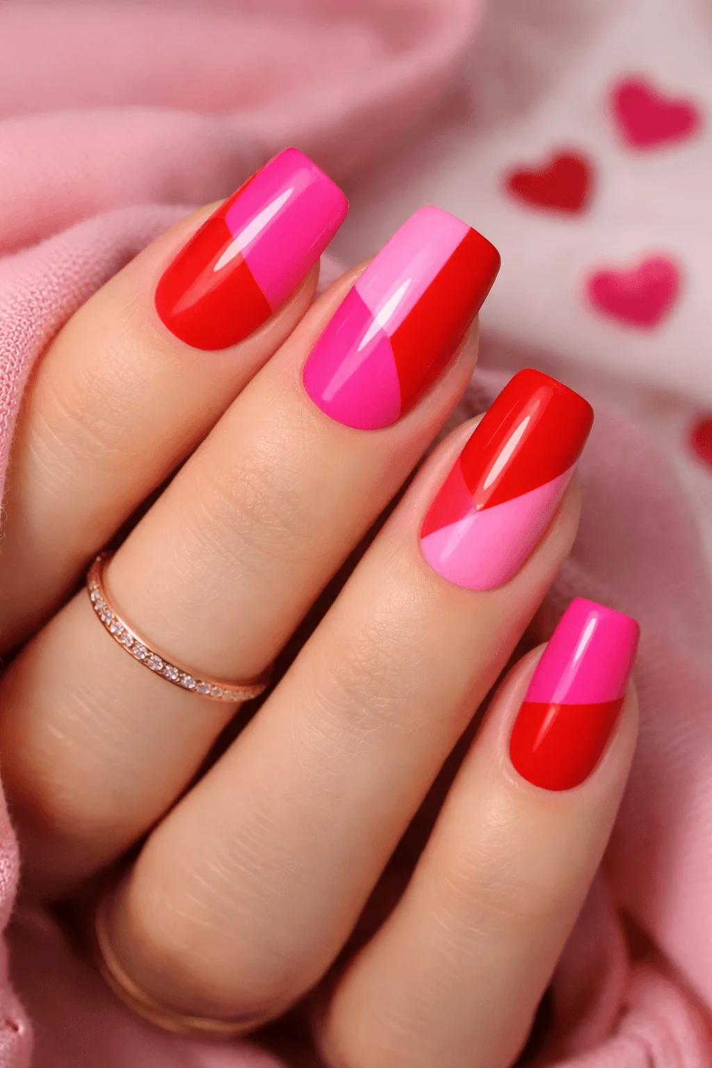 Close up of a hand with glossy pink and red color block nails featuring diagonal and half and half designs and a soft pink background