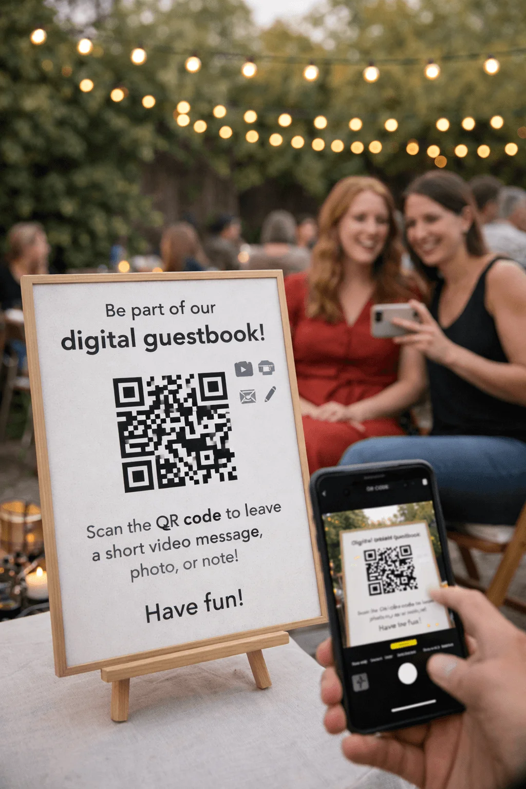 Framed sign on a small wooden easel reading “Be part of our digital guestbook!” with a large QR code and instructions to scan it to leave a short video message photo or note while a guest holds a smartphone scanning the code and two women smile in the softly lit outdoor party setting with string lights in the background