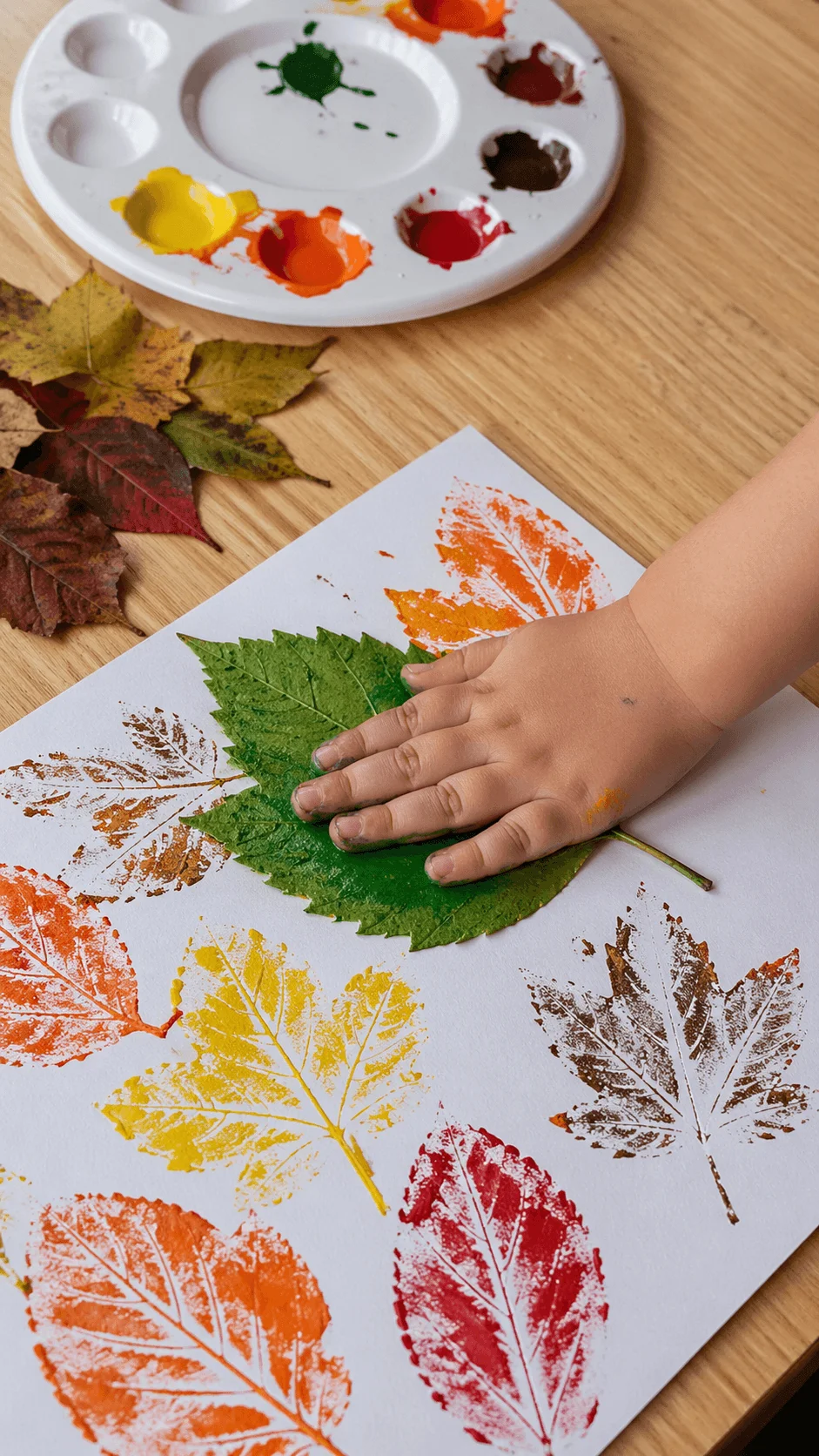 Toddler making leaf prints by pressing painted leaves onto paper with colorful paint palette nearby