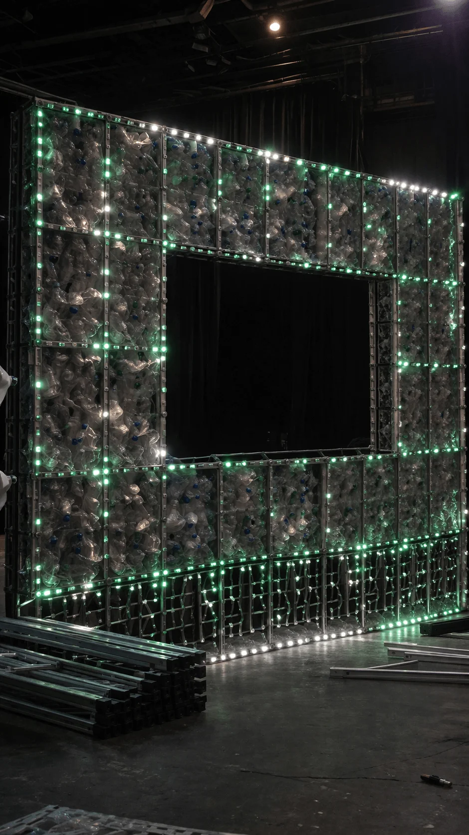 Stage structure made from recycled plastic bottles arranged in modular panels with embedded LED lights forming a rectangular frame with an open center in a workshop setting