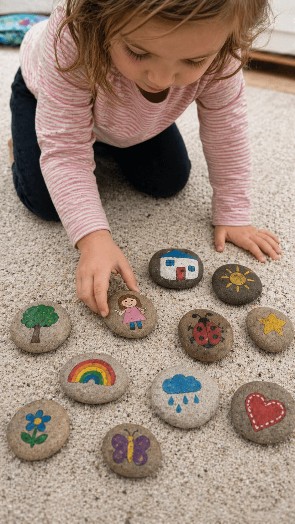 Child arranging painted story stones with colorful drawings on a carpet floor