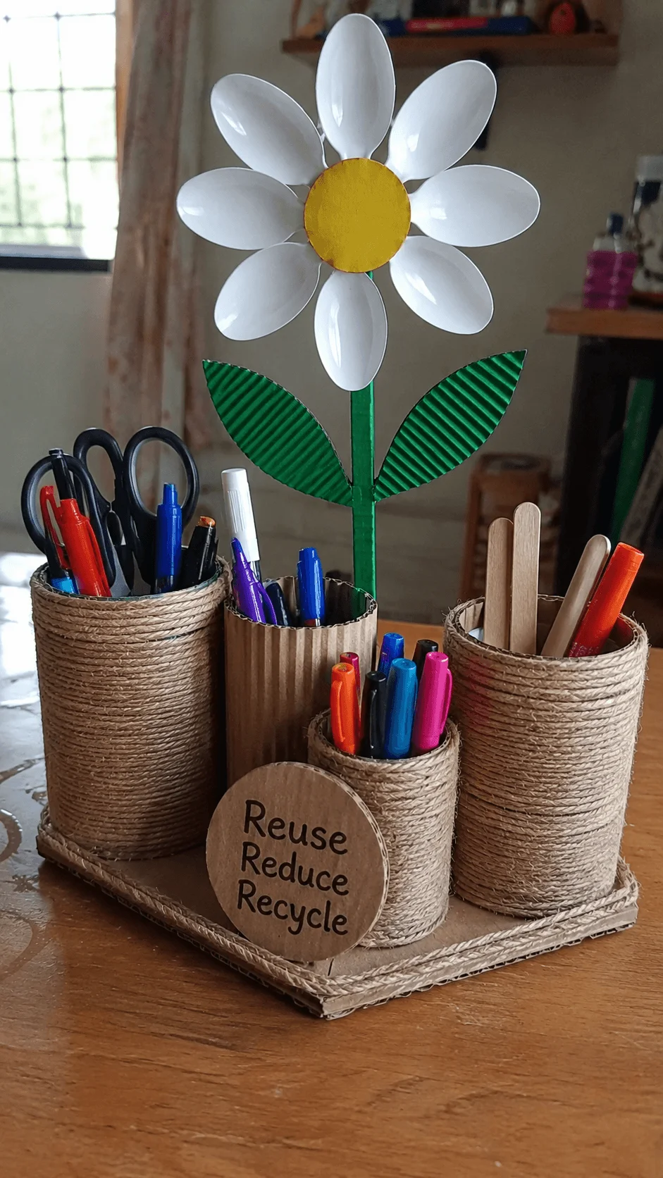 DIY recycled desk organizer made from cardboard tubes and rope with pens scissors and craft sticks arranged in separate holders topped with a flower made from plastic spoons and a sign reading Reuse Reduce Recycle