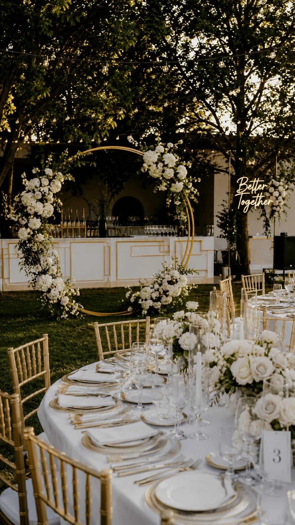 Elegant outdoor wedding reception with round tables white floral arrangements gold chairs and a circular flower arch under string lights in a garden setting at sunset