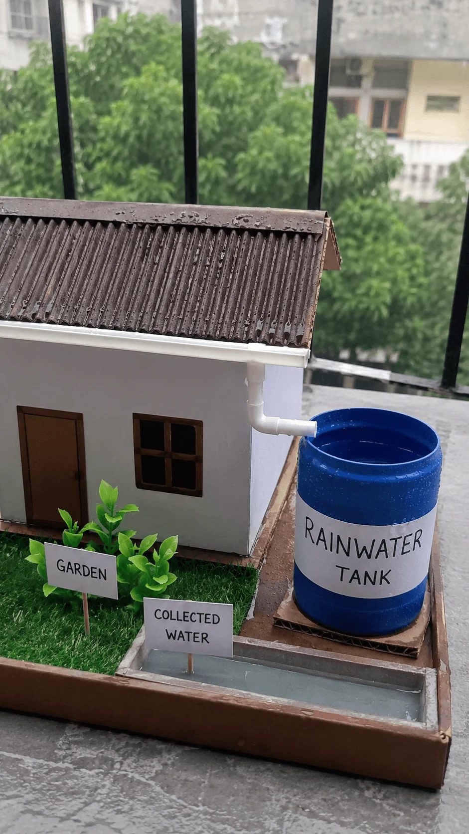 Model house demonstrating an easy rainwater harvesting project for kids with rooftop drainage pipe water collection tank garden area and stored rainwater system for home sustainability learning