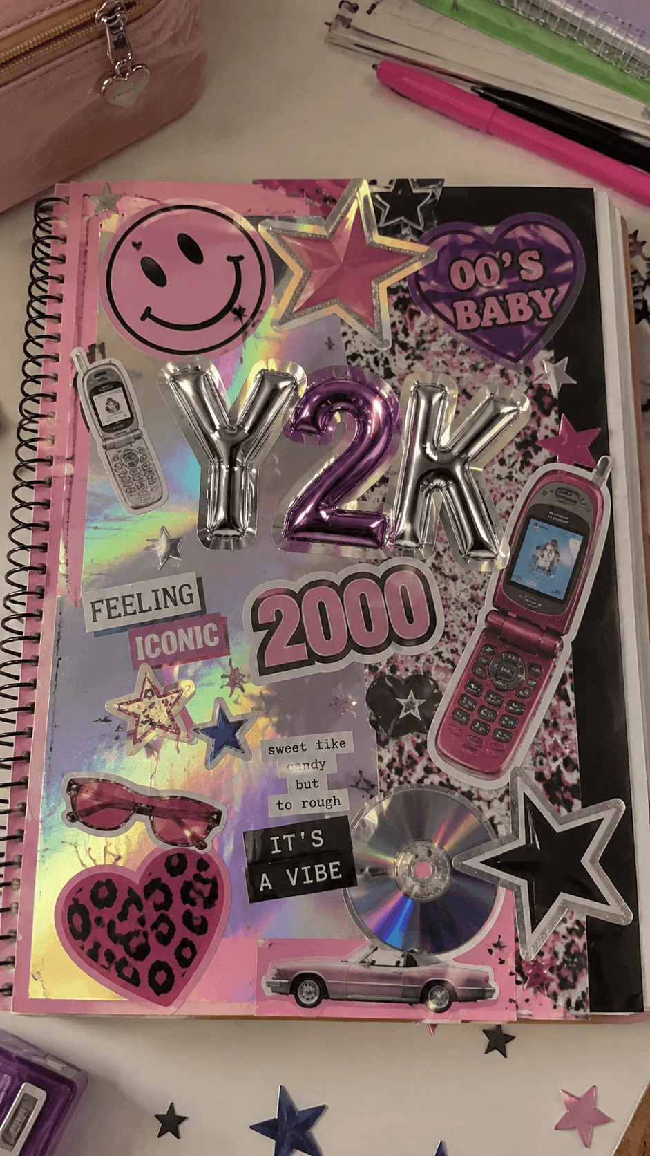Retro Y2K aesthetic scrapbook cover featuring holographic textures flip phones smiley faces stars pink and purple stickers metallic balloon letters and nostalgic early 2000s fashion elements arranged on a spiral notebook with colorful stationery on a desk