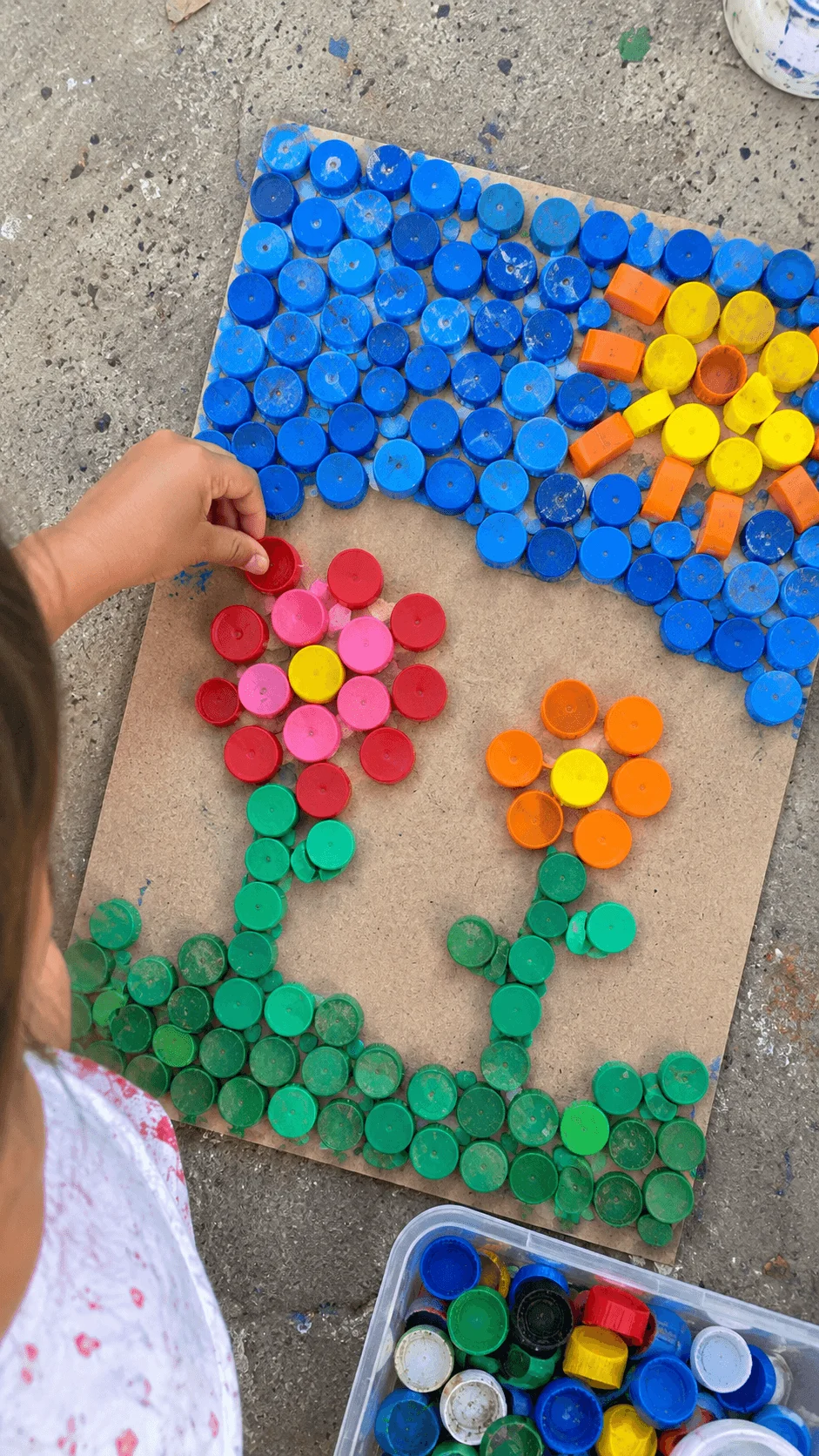Child creating a colorful mosaic artwork from recycled plastic bottle caps on a wooden board, arranging flower shapes beneath a bright sun and blue sky design outdoors.