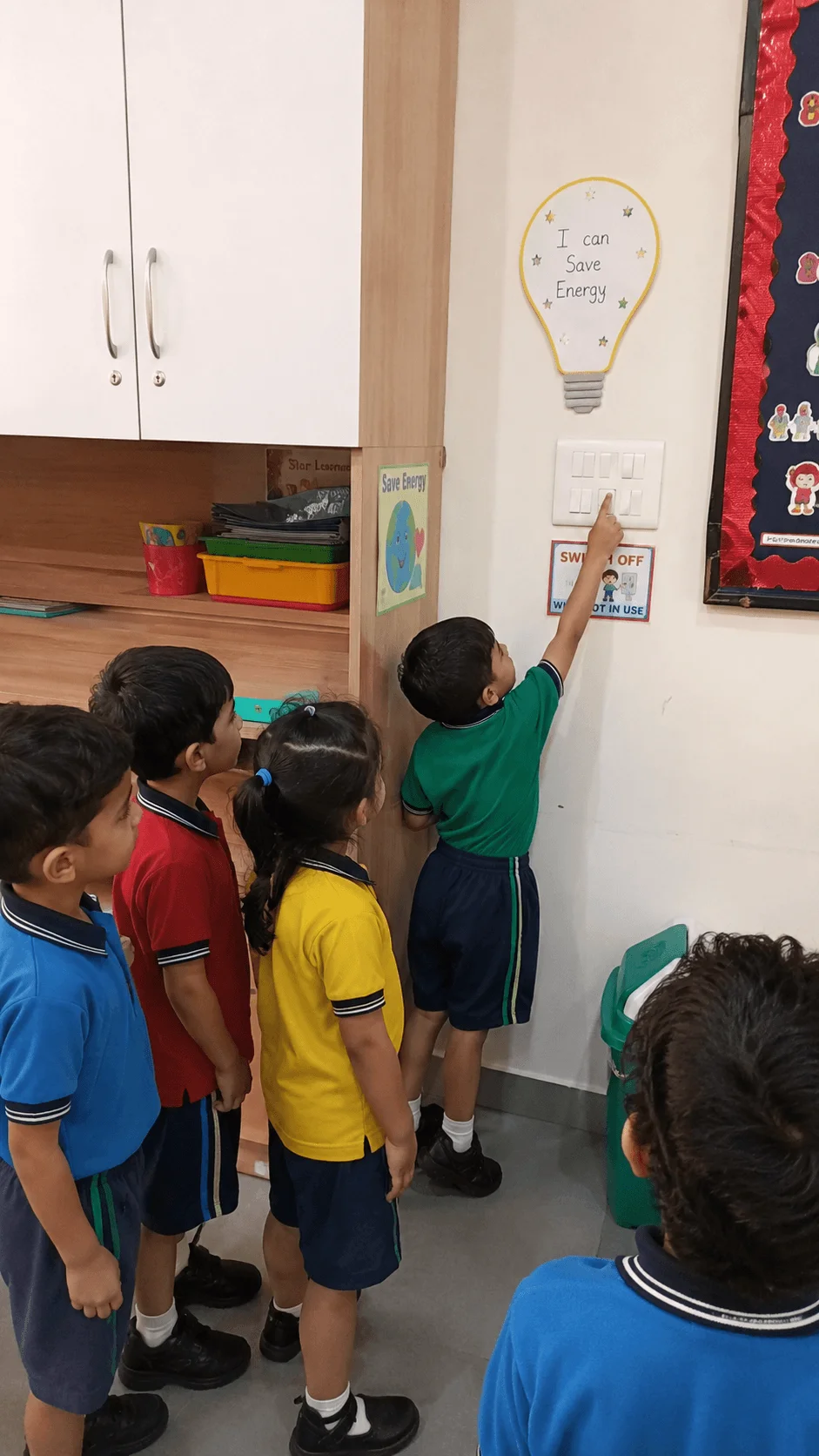 Preschool children taking turns switching off classroom lights during an energy saving role play activity that teaches responsible daily habits.