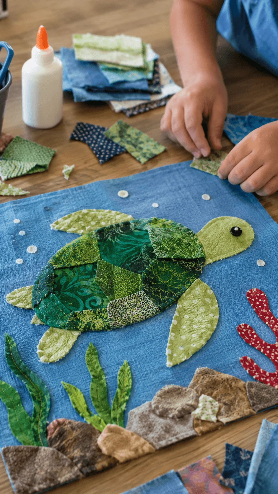 Fabric scrap animal art project featuring a sea turtle created from colorful fabric pieces on a blue background with underwater plants coral and rocks while a child assembles the eco friendly collage using recycled textiles.