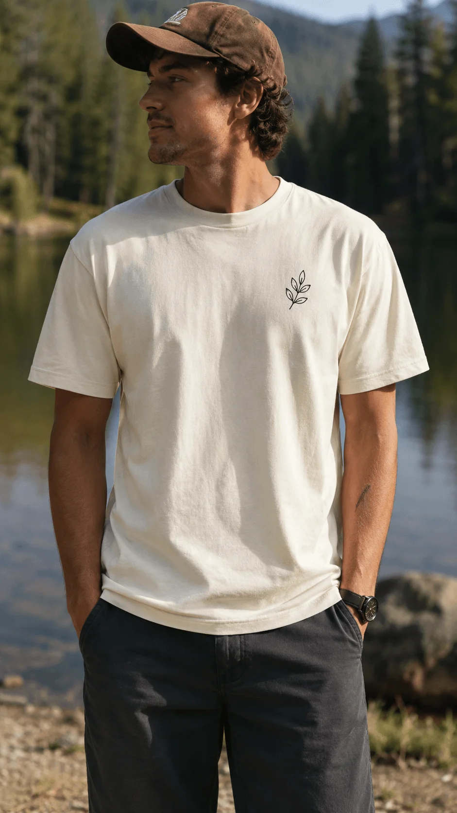 Man wearing a minimalist sustainable white T shirt with a small leaf design beside a mountain lake