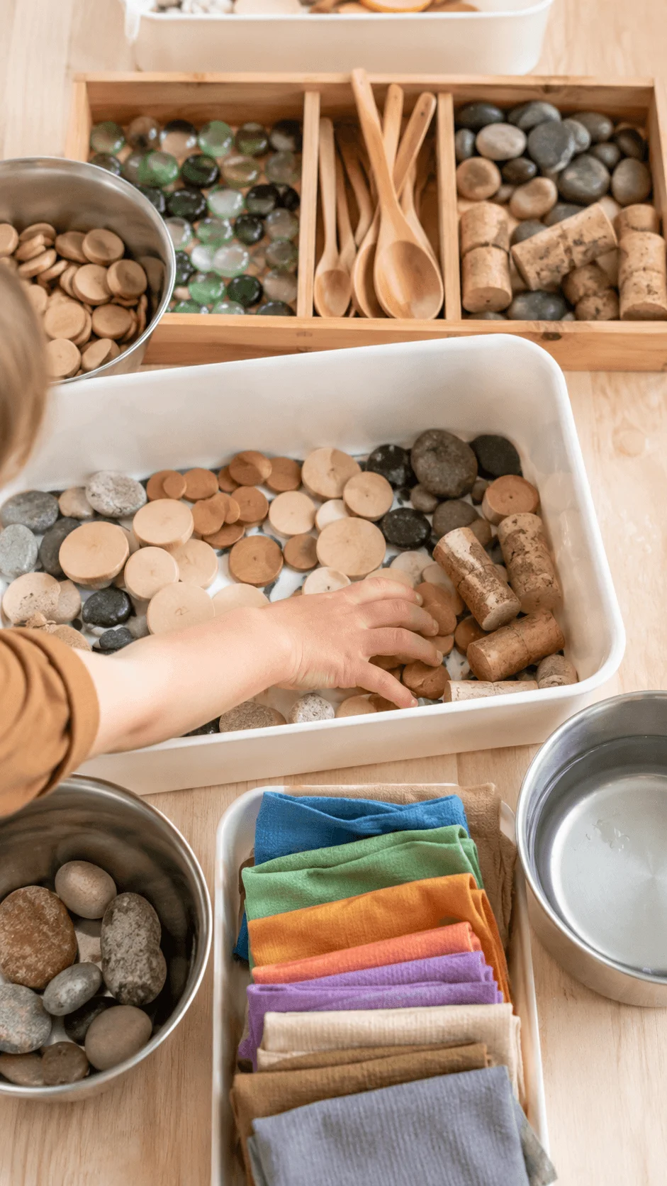 Child exploring low waste sensory play with reusable natural materials including wooden discs stones corks fabric pieces and wooden spoons in a sustainable learning activity