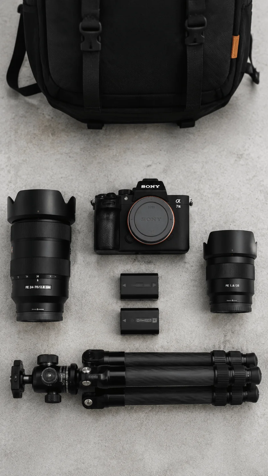 Minimalist photography gear setup featuring a mirrorless camera, lenses, batteries, tripod, and camera bag arranged neatly on a clean surface for professional photography use