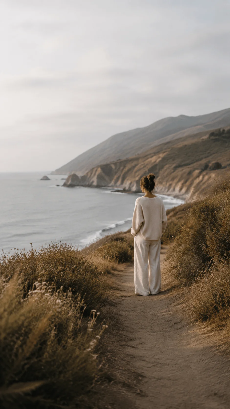 Person standing on a coastal trail overlooking the ocean and mountains with soft natural light and low contrast photography editing style