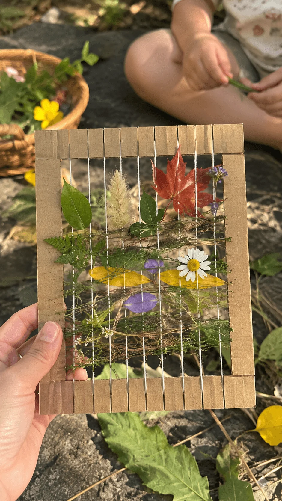 Nature weaving craft made with a cardboard loom and natural materials including leaves flowers grass and plant stems during an outdoor creative exploration activity for kids.