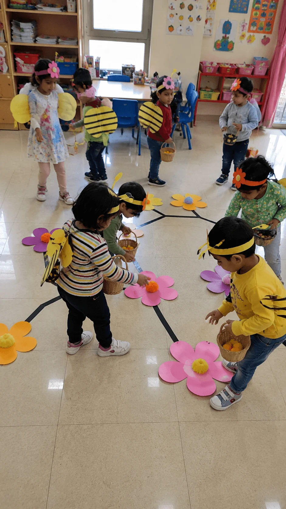 Preschool children dressed as bees and flowers moving between colorful flower props during a classroom pollinator awareness game about nature connections.