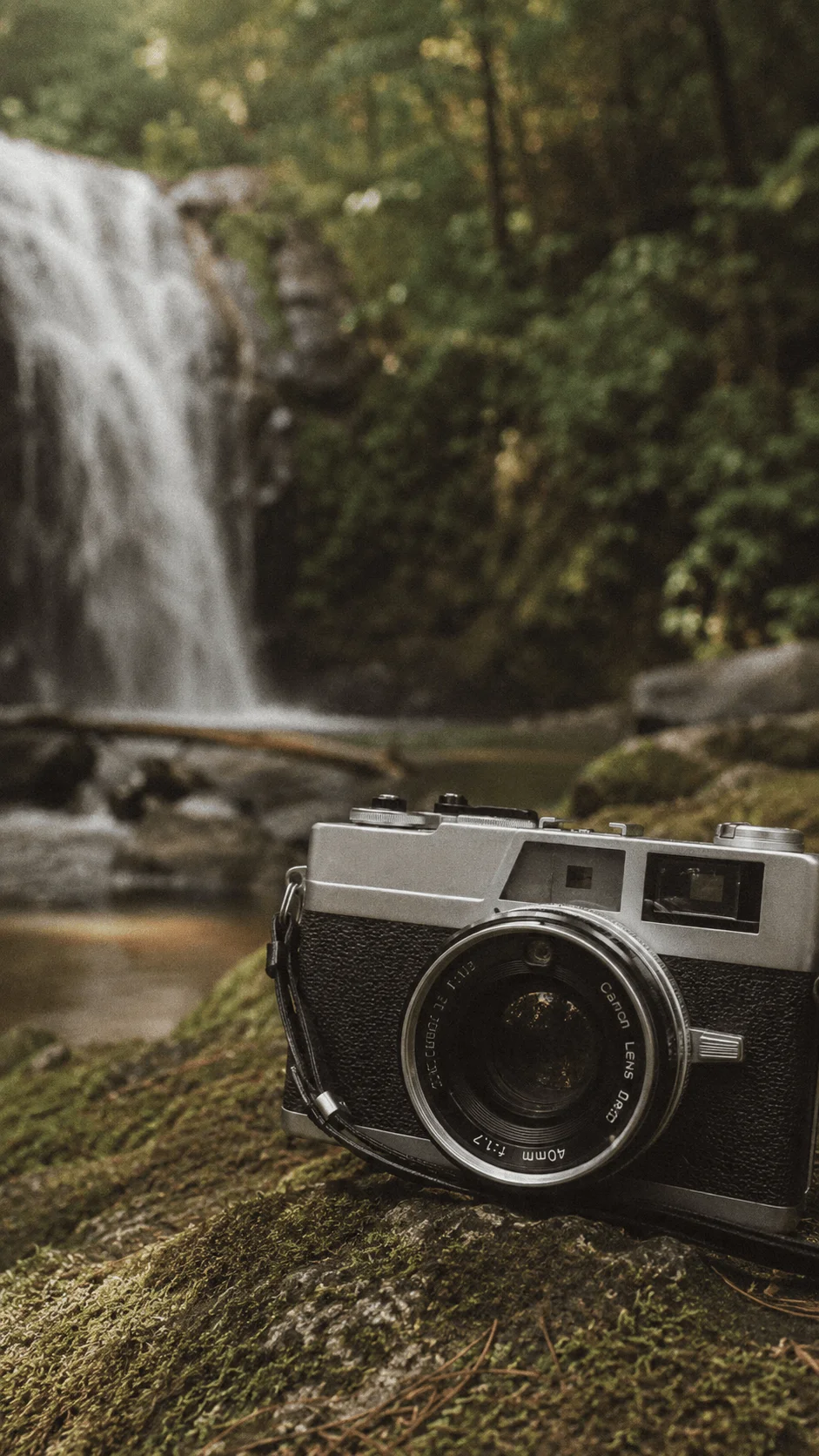 Vintage film camera resting on a moss covered rock beside a forest waterfall surrounded by lush greenery