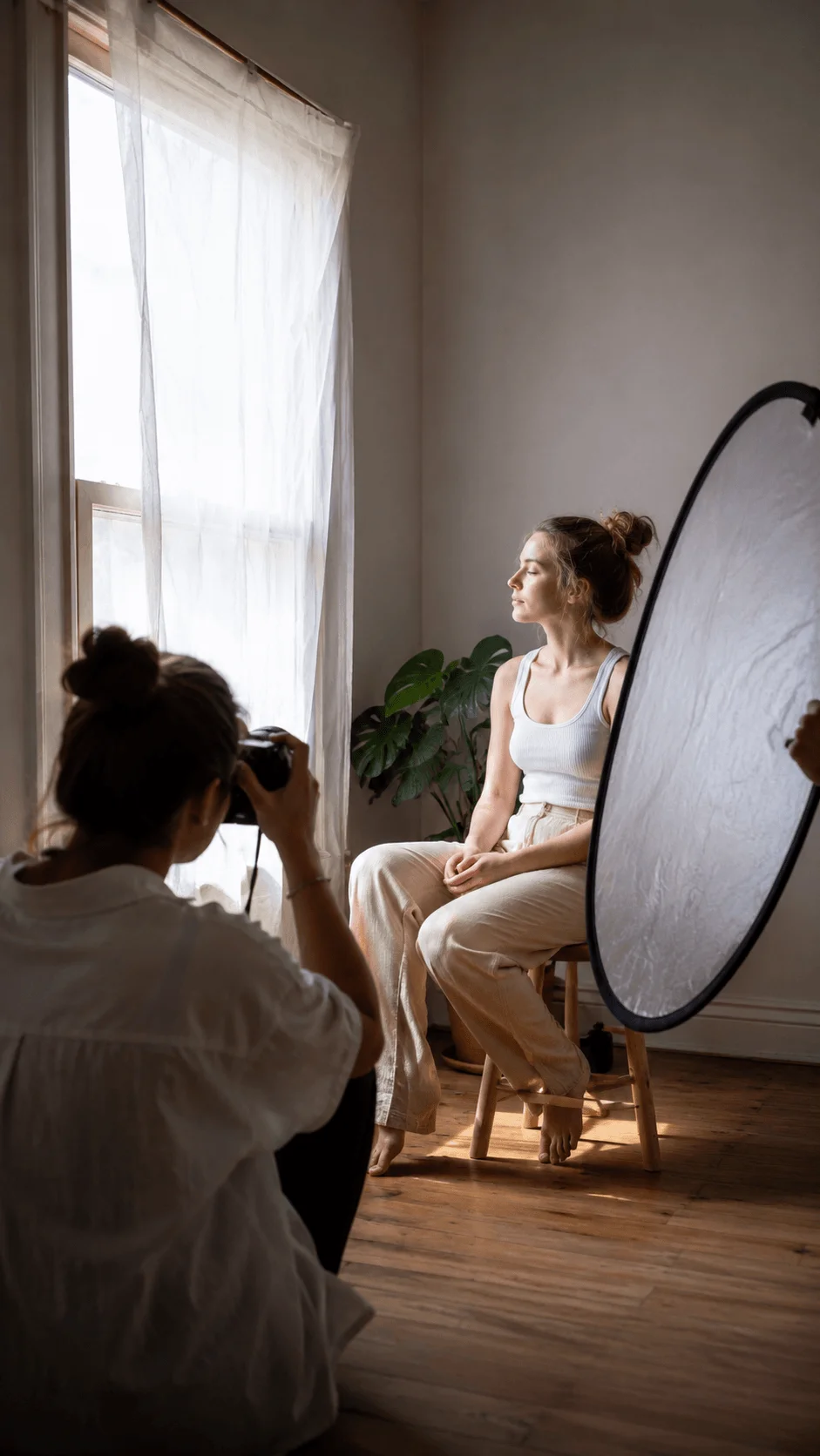 DIY window diffusion setup for natural photography lighting with a photographer capturing a seated subject beside a sheer curtain, soft window light, and a large reflector in a warm minimalist studio