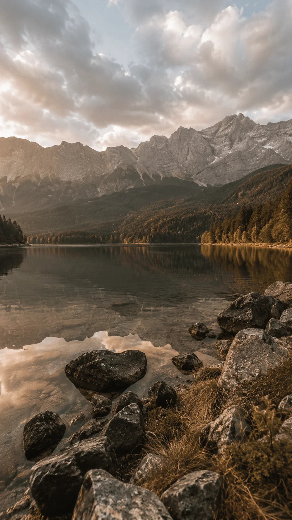 Earth tone mountain landscape with a calm reflective lake rocky shoreline evergreen forest and dramatic cloudy sky