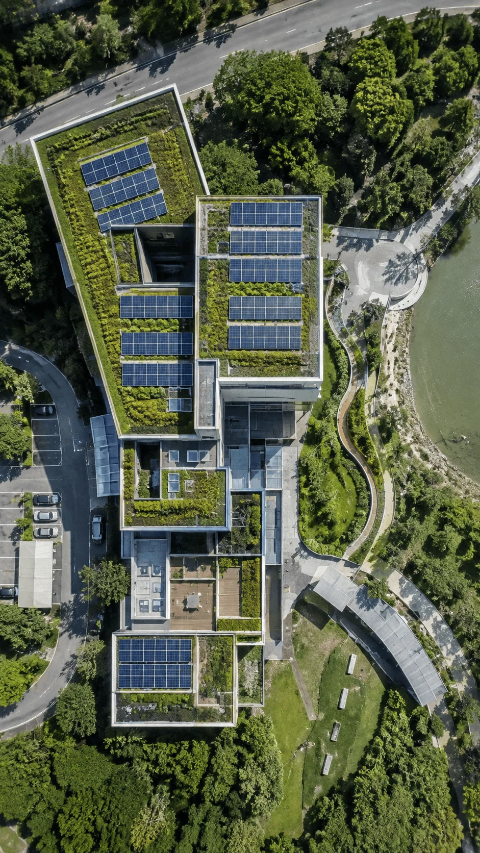 Aerial view of a modern eco friendly building with green rooftops and solar panels surrounded by trees, roads, and a waterfront park.