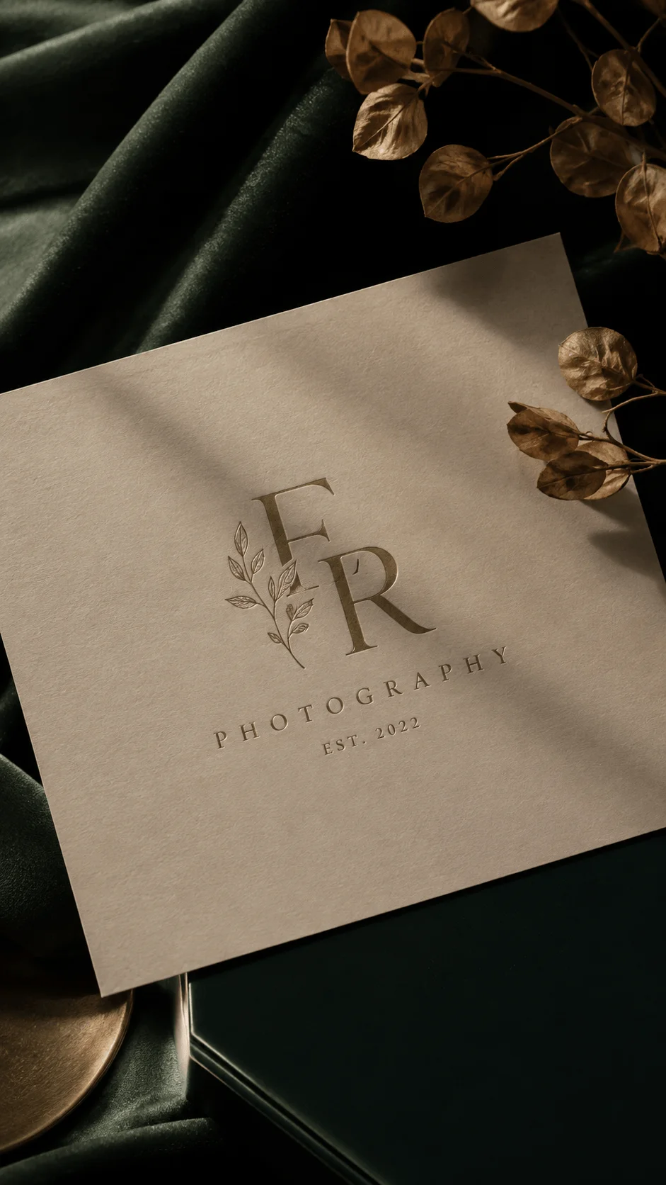 Eco luxury photography logo featuring elegant serif initials with a botanical leaf accent on textured paper surrounded by dried leaves and dark natural fabric background