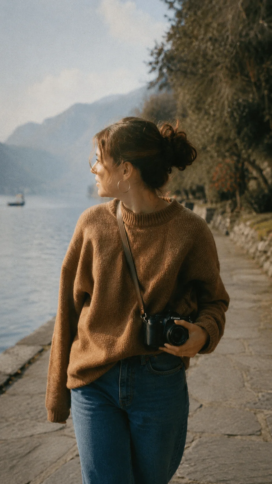 Woman holding a camera while walking beside a peaceful mountain lake in warm sunlight