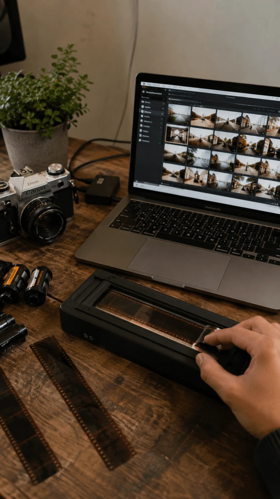 Hybrid analog and digital photography setup with a Canon film camera, laptop displaying photos, film scanner and photographic negatives on a wooden desk