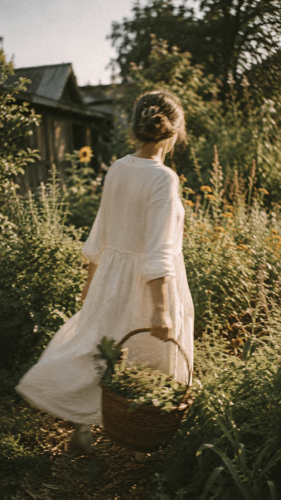 Woman in a white dress carrying a woven basket while walking through a sunlit garden surrounded by plants and flowers