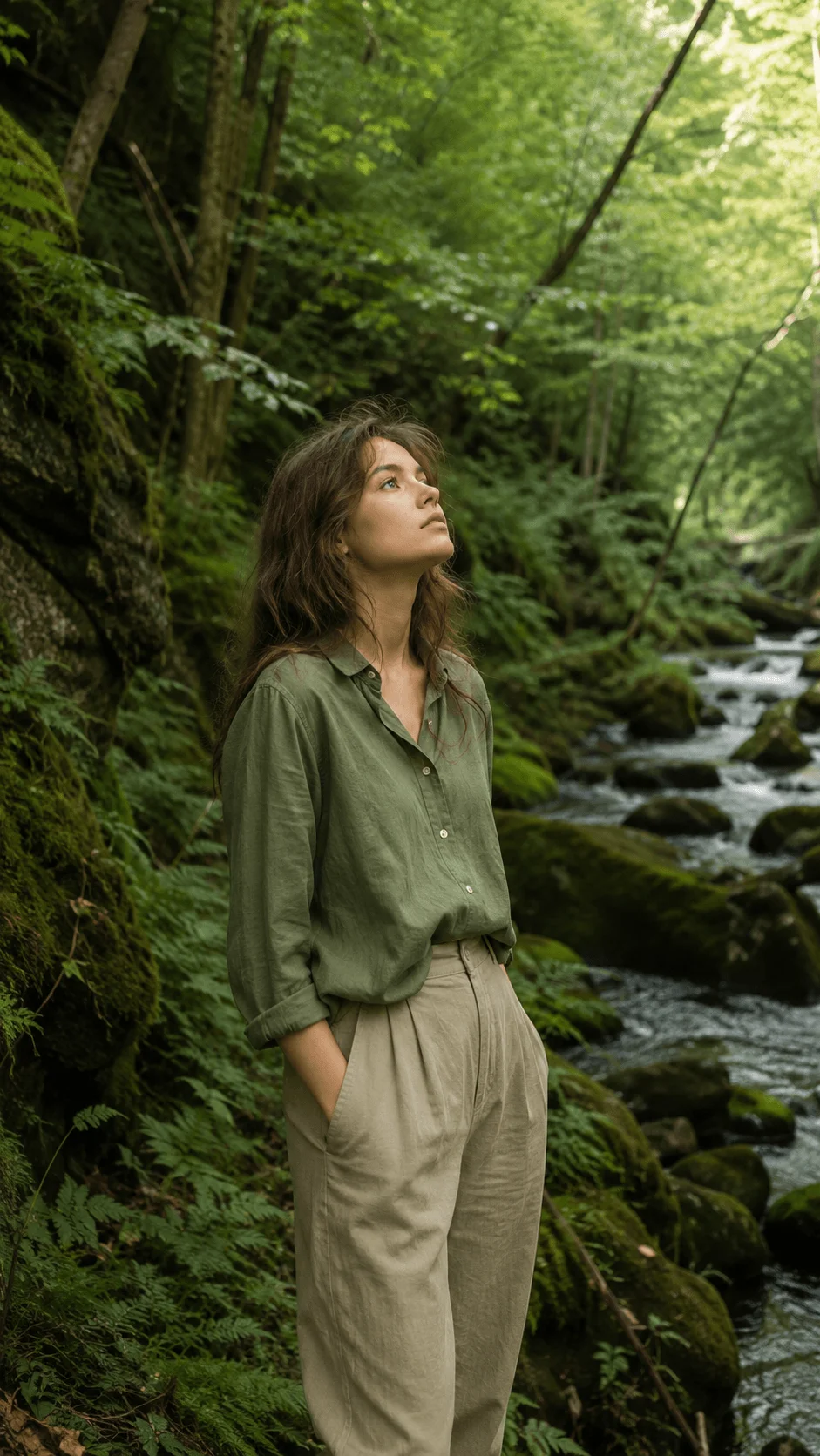 Woman standing beside a forest stream surrounded by lush green trees and moss covered rocks