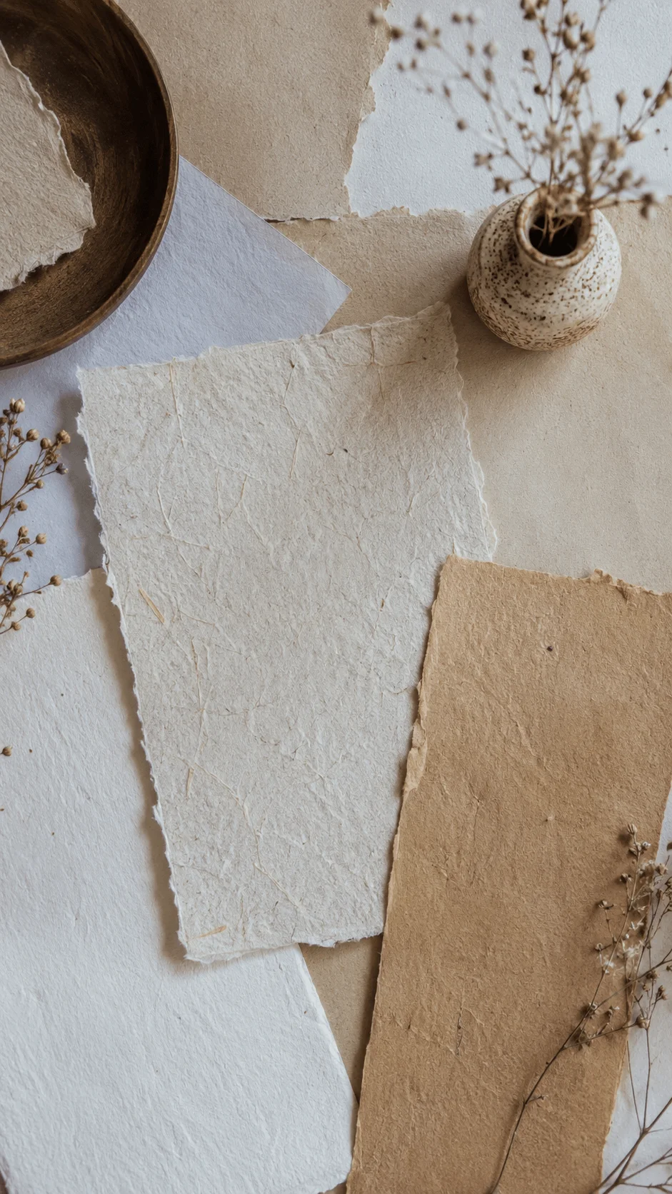 Recycled and handmade paper photography backgrounds with textured neutral paper sheets, dried flowers, and a small ceramic vase arranged in a natural minimalist setting