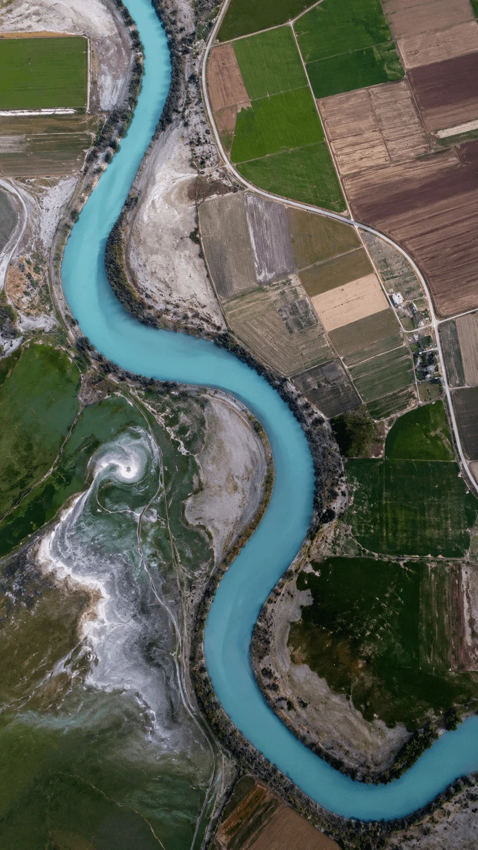 Aerial view of a bright turquoise river winding through agricultural fields, exposed rocky terrain, green farmland, and winding roads.