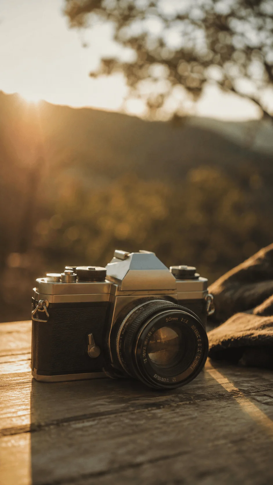 Vintage camera with a classic lens resting on a wooden table in warm golden sunlight with soft mountains and trees in the background
