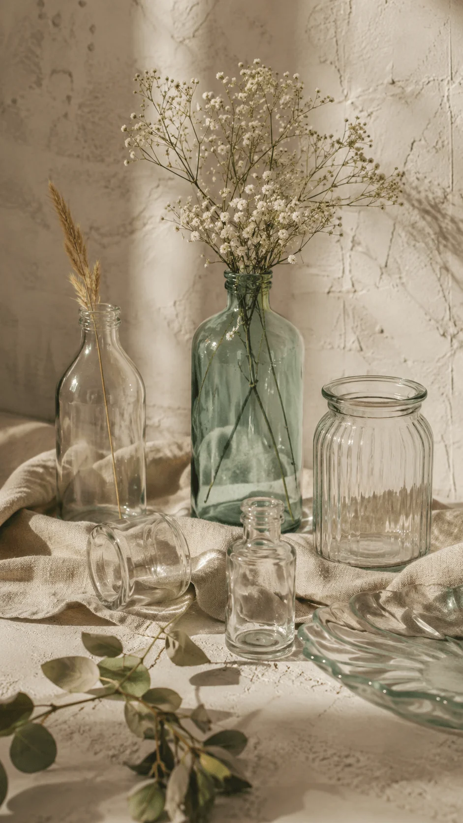 Upcycled glass photography props with recycled glass bottles, jars, dried flowers, linen fabric, eucalyptus leaves, and textured neutral surfaces in warm natural light