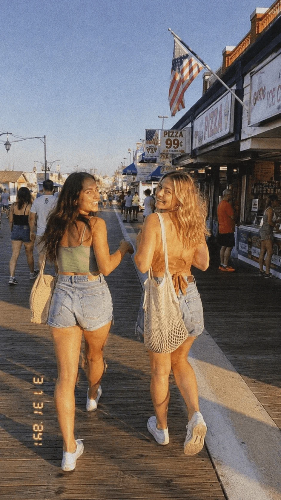 Two women walking along a lively beach boardwalk in casual summer outfits with tote bags during golden hour.