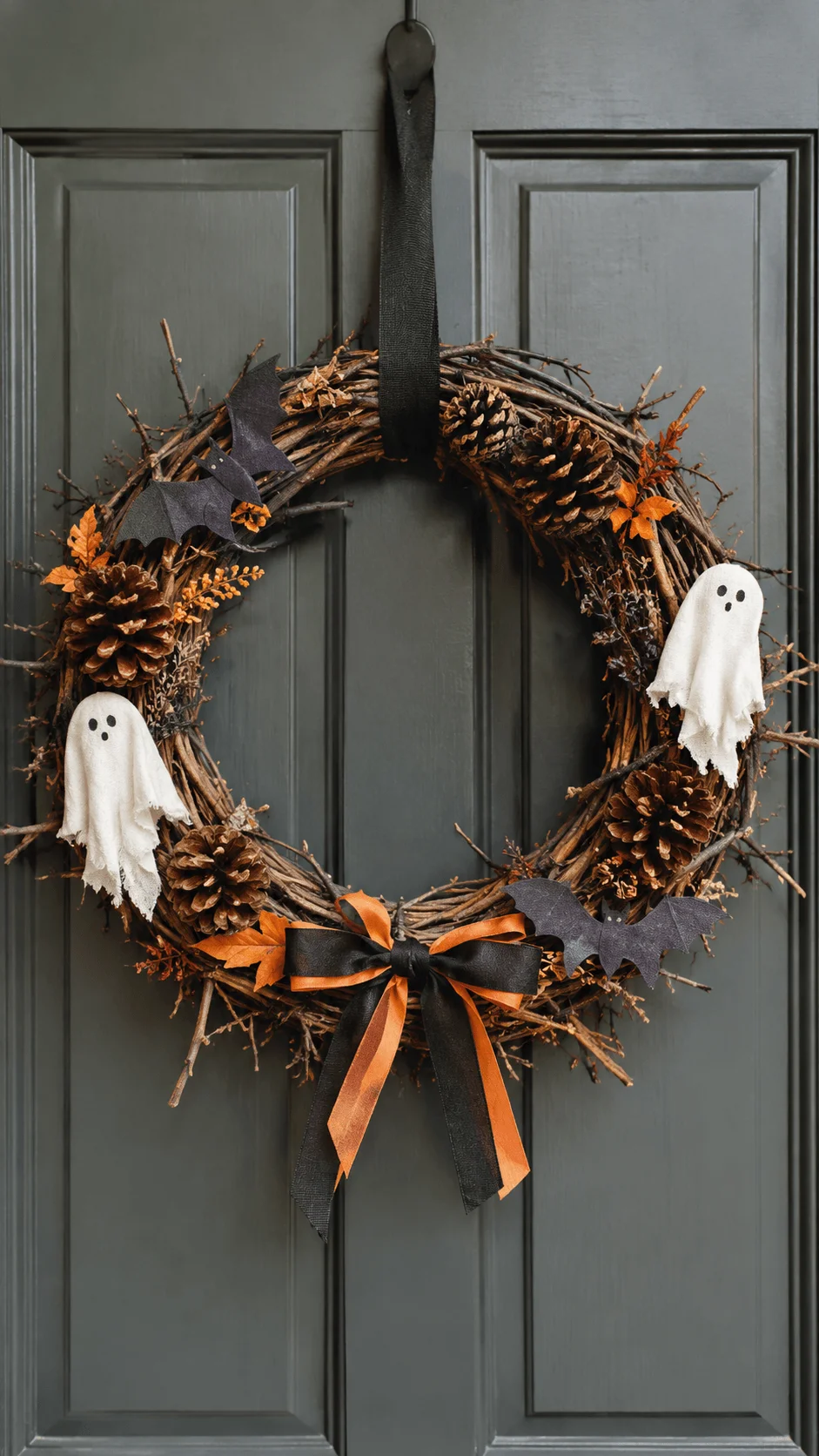 Rustic Halloween wreath made from natural twigs and pinecones, decorated with white fabric ghosts, black bats, dried orange foliage and a black and orange ribbon bow hanging on a gray front door for eco friendly Halloween decor